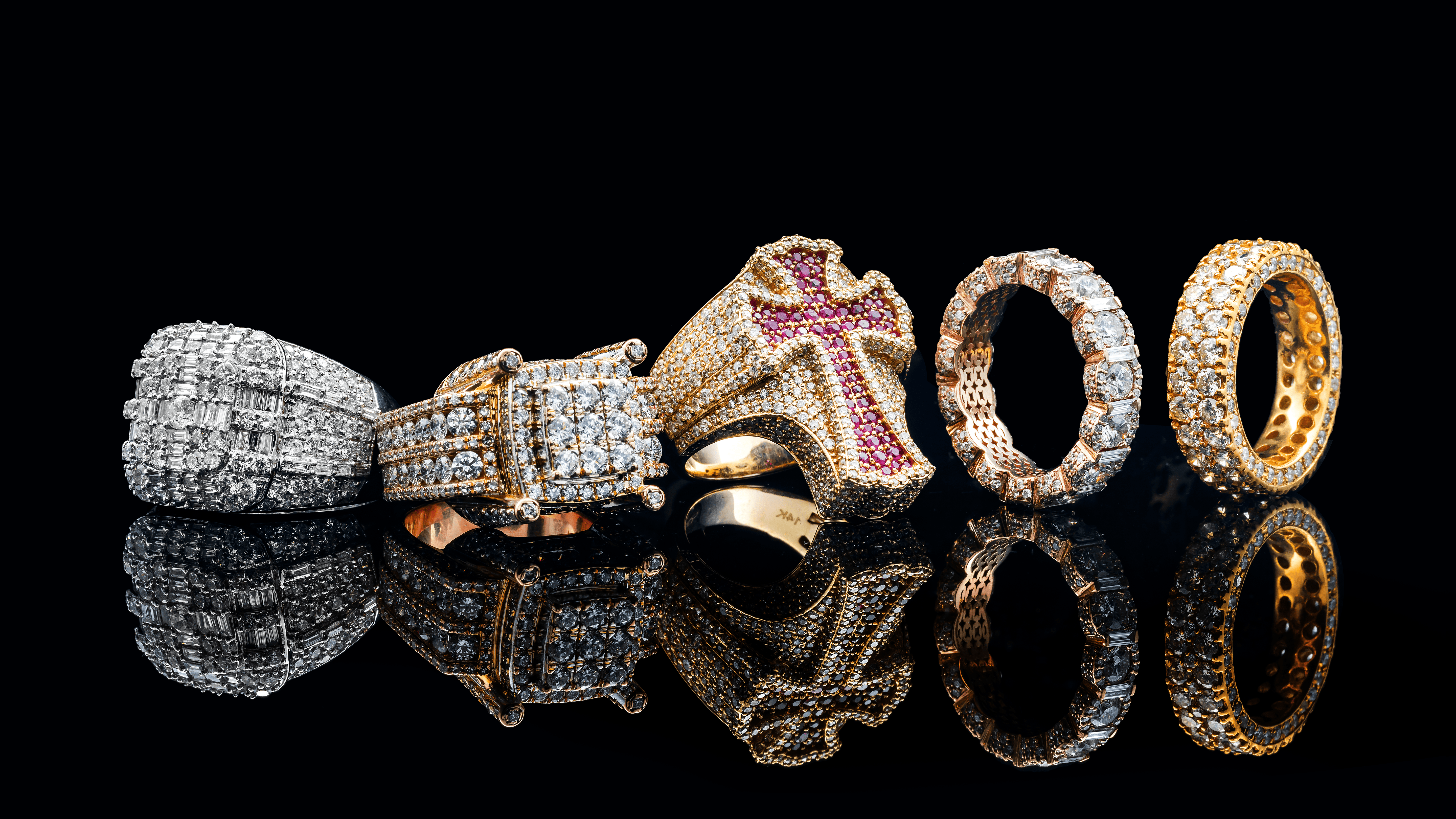A lineup of six luxurious men's diamond rings displayed against a glossy black background. The rings feature various designs, including pavé and baguette diamond settings in white gold, rose gold, and yellow gold. One standout ring in the center is a bold gold piece with a red ruby cross design, while the others vary in width and diamond arrangement, showcasing a mix of modern and bold aesthetics with brilliant sparkle and detailed craftsmanship